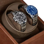 Close up of two watches, one with a black face and silver band, the other with a blue face and silver band, in a brown watch winder box.