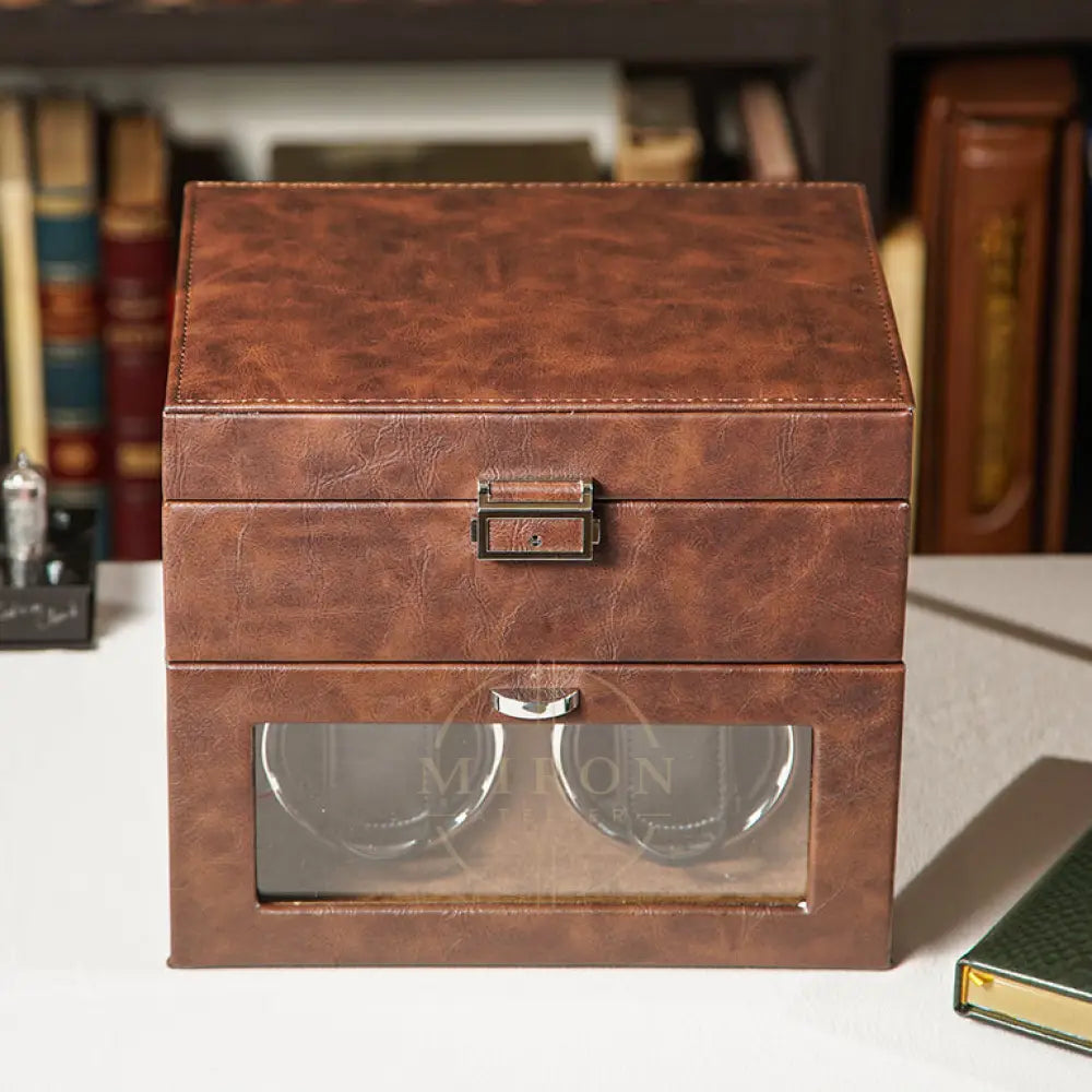 Brown leather watch box with glass lid on a surface with books in the background