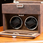 Brown leather watch winder box with two rotating watch cases on a wooden surface.