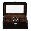 A luxury watch wooden box with a dark wood grain exterior, a clear glass lid, and a brown velvet interior. A silver and black chronograph watch is displayed in one of the three watch slots.