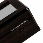 Close-up of a dark wood watch box with a glossy finish, showing a glass window and silver metal hinge details on a white background.