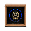 A luxury watch winder featuring a medium brown wood-grain frame and a black interior with a glowing blue circular light. A gold and black chronograph watch is displayed inside the center.