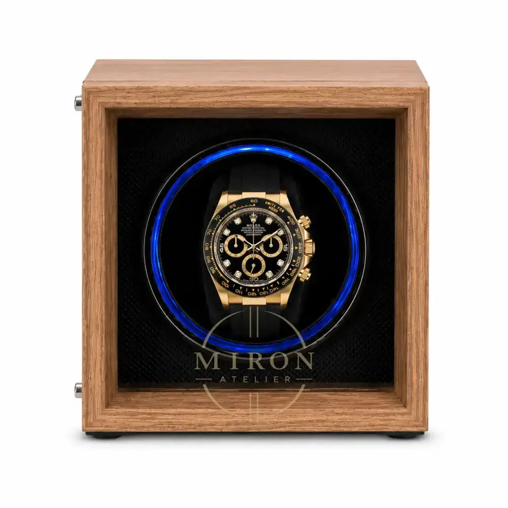 A luxury watch winder featuring a medium brown wood-grain frame and a black interior with a glowing blue circular light. A gold and black chronograph watch is displayed inside the center.
