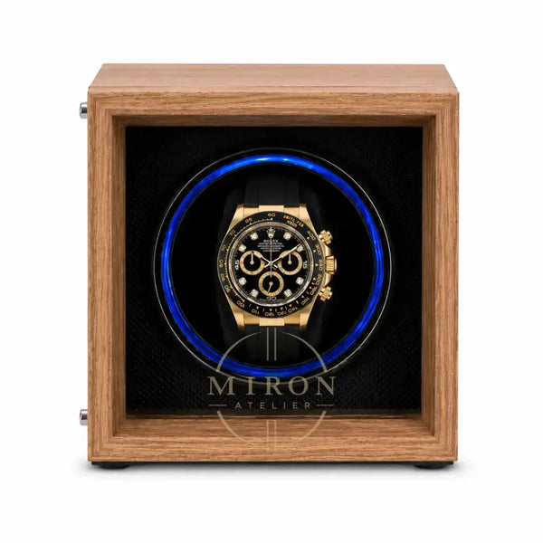 A luxury watch winder featuring a medium brown wood-grain frame and a black interior with a glowing blue circular light. A gold and black chronograph watch is displayed inside the center.