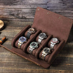 A brown leather watch roll with six slots holding six watches, photographed on a rustic wooden tabletop.
