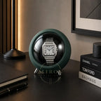 Cartier watch in a green watch winder on a desk with books and a lamp.
