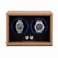 A wooden dual watch winder with a medium brown wood-grain finish, black textured interior, and two circular watch slots illuminated by blue LED lights. Two stainless steel watch dials are displayed inside as demonstration props.